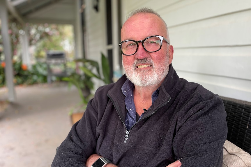 A man with short grey hair and glasses wearing a jumper sits on a seat outside, smiling.