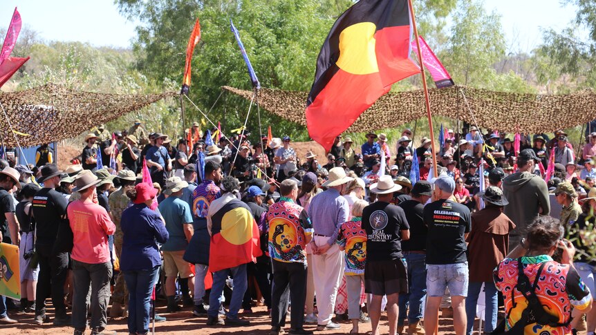 A crowd of people holding Aboriginal flags in a bush setting, on a sunny day.