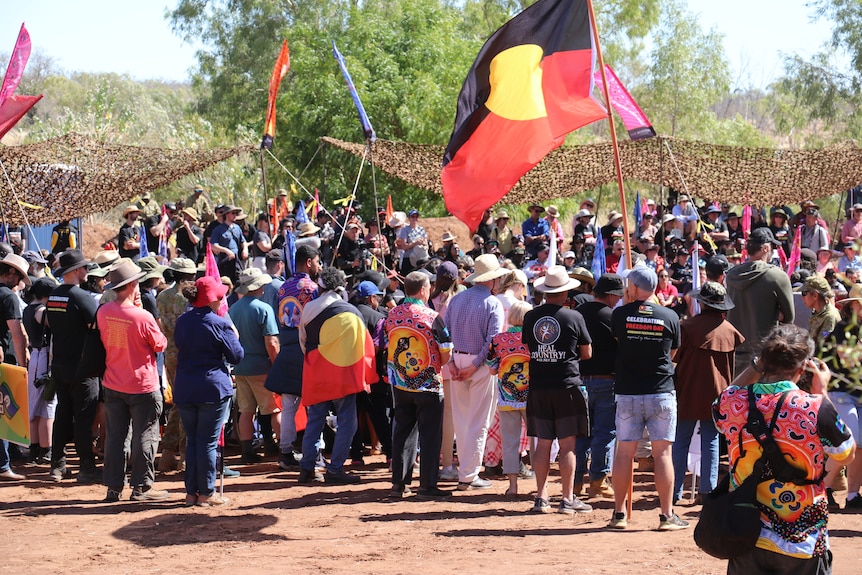 A crowd of people holding Aboriginal flags in a bush setting, on a sunny day.