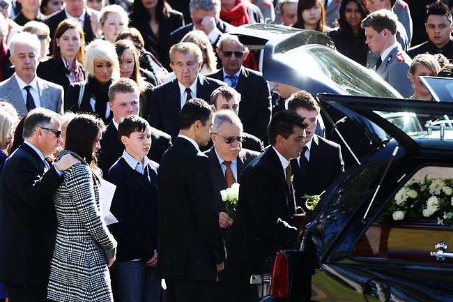 People gather around a hearse as a coffin is placed in the back.