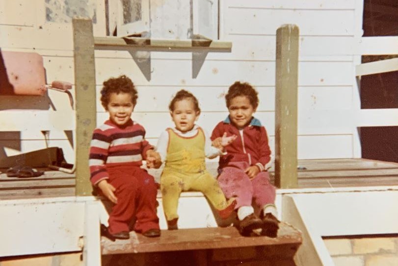A toddler girl sits between her two brothers on a set of steps of a home