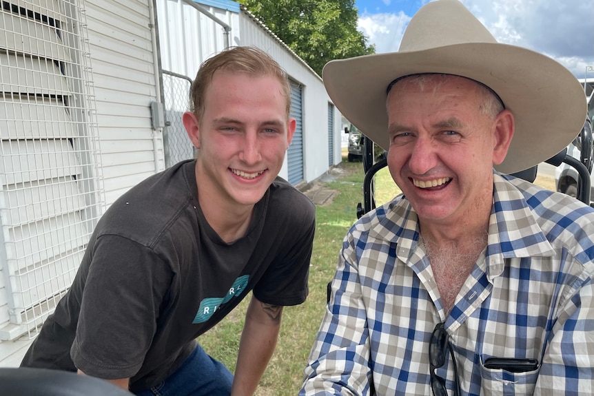 A man in a check shirt and a cowboy hat smiles. He is next to another younger man in a black shirt.