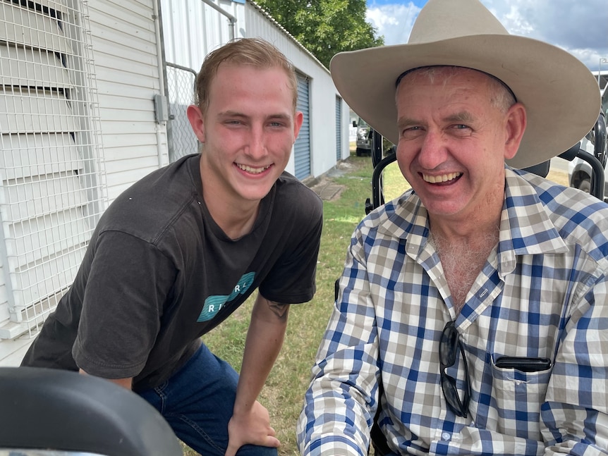 A man in a check shirt and a cowboy hat smiles. He is next to another younger man in a black shirt.