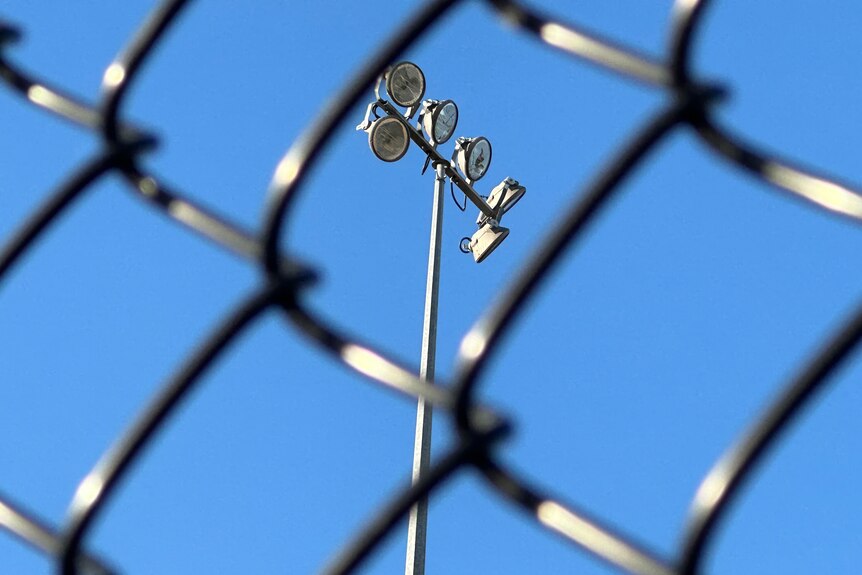 Large sports field flood lights, viewed through a fence on a clear day.