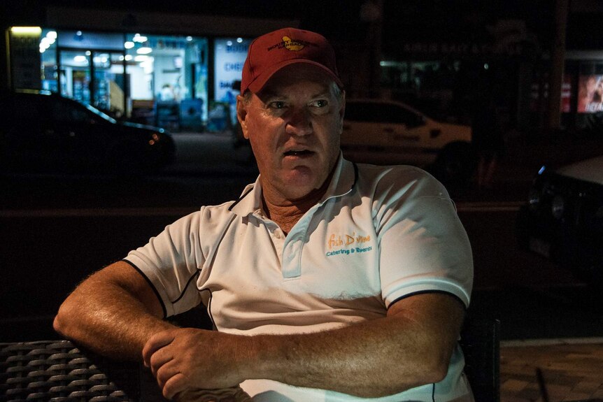Business owner Kevin Collins sits on a bench at night looking unhappy, with shops and cars behind him.