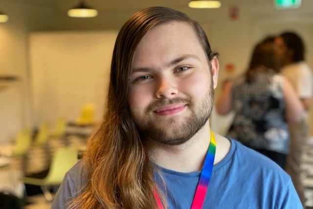 Avery Howard smiles and looks into the camera. He has long hair and wears a rainbow lanyard over a blue shirt.