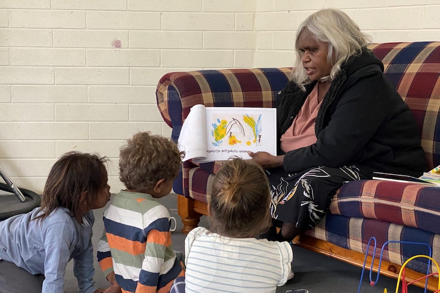 An Aboriginal woman sitting on a sofa reads a book in an Indigenous language to children sitting on the floor