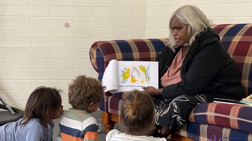 An Aboriginal woman sitting on a sofa reads a book in an Indigenous language to children sitting on the floor