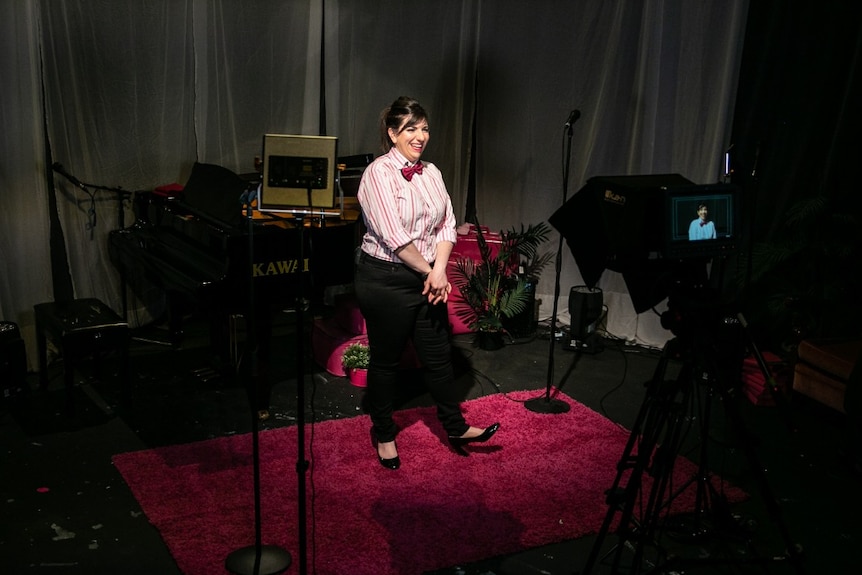 A woman wearing a shirt and bow tie smiles into a camera set up with lighting in a studio.