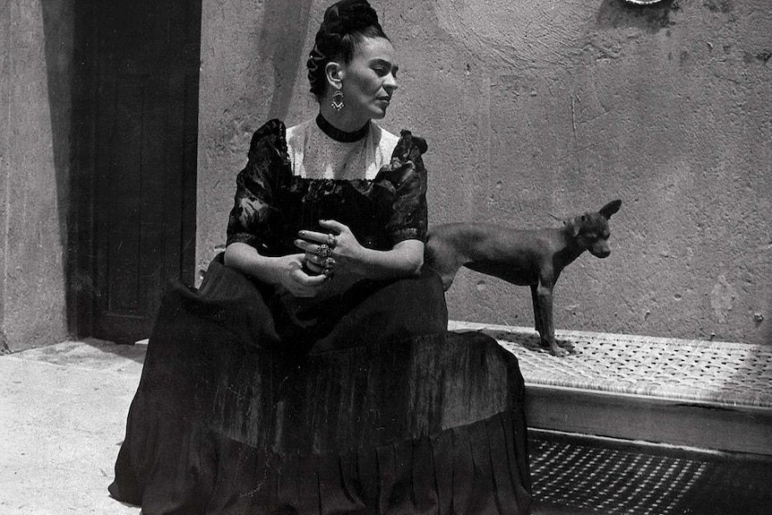 A black and white photo of Frida Kahlo with traditional Mexican dress and hair, sitting on a day bed in a courtyard with a dog.