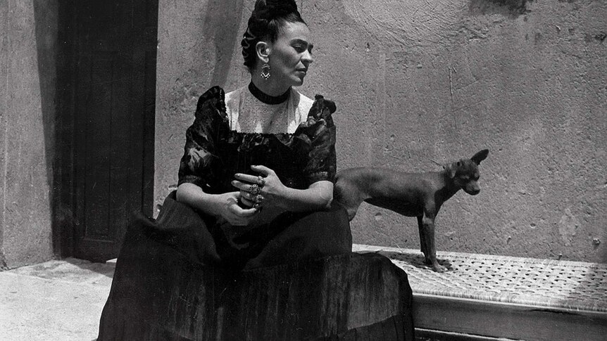 A black and white photo of Frida Kahlo with traditional Mexican dress and hair, sitting on a day bed in a courtyard with a dog.