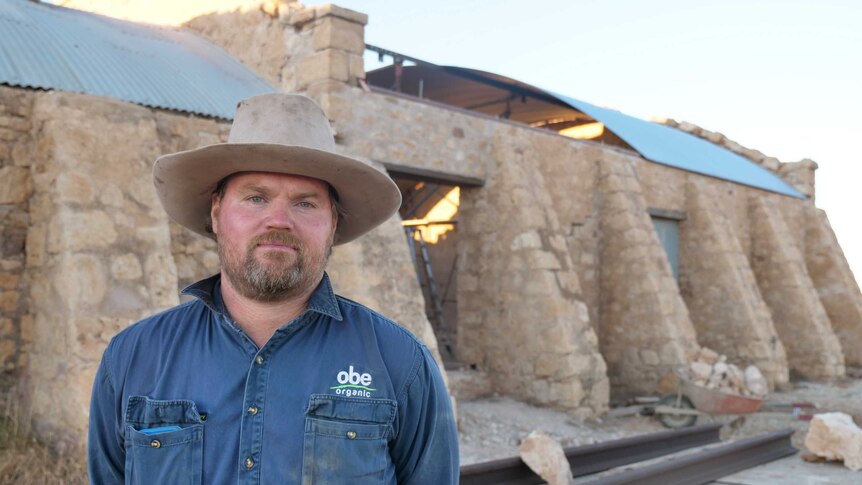 A man in a dark blue long sleeve shirt and light brown wide-brimmed hat stands in front of the stonework of the woolshed.