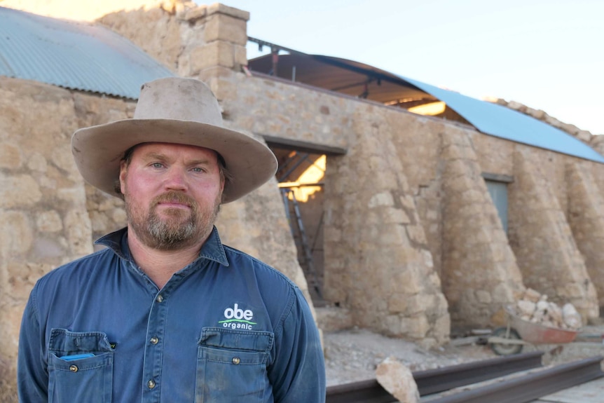 A man in a dark blue long sleeve shirt and light brown wide-brimmed hat stands in front of the stonework of the woolshed.