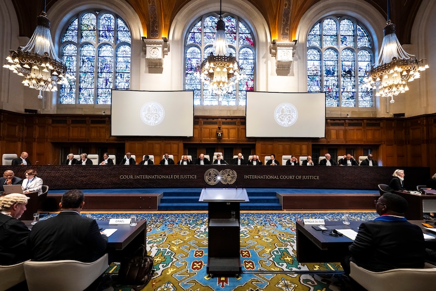 In room with high wooden ceiling and huge stained glass windows, 17 judges wearing black sit behind a wooden bench.