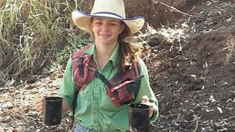 Dolly Everett, wearing a wide-brimmed hat and carrying two mugs, smiles as she walks in the bush.