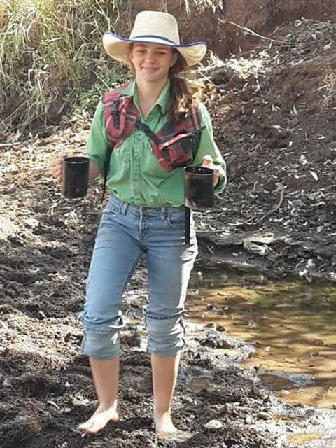 Dolly Everett, wearing a wide-brimmed hat and carrying two mugs, smiles as she walks in the bush.