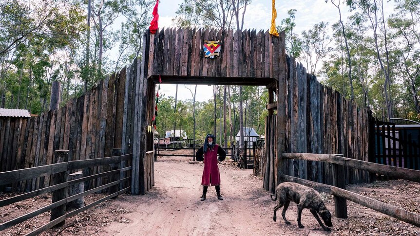 A man stands in  medieval clothing outside gates made of timber and a large shaggy dog.