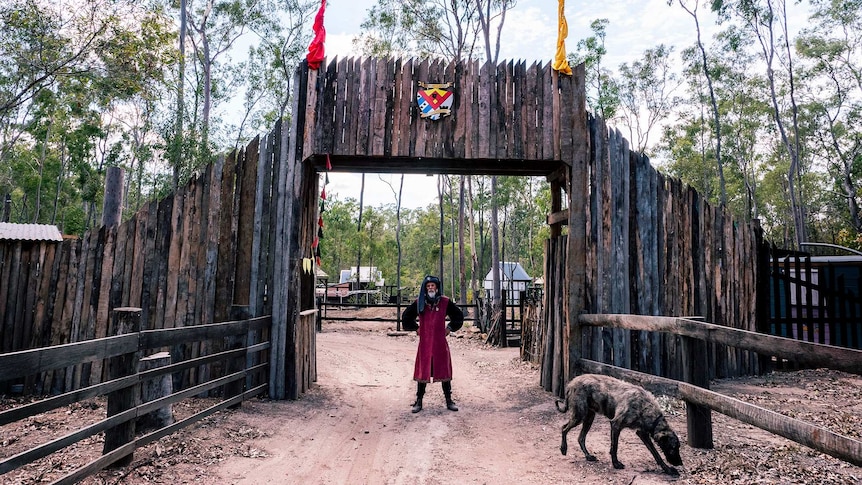 A man stands in  medieval clothing outside gates made of timber and a large shaggy dog.