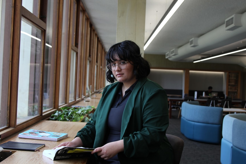 Serious girl, black bob, glasses, green jacket, blue top, holds book, looks at camera, in room with windows, blue couches.