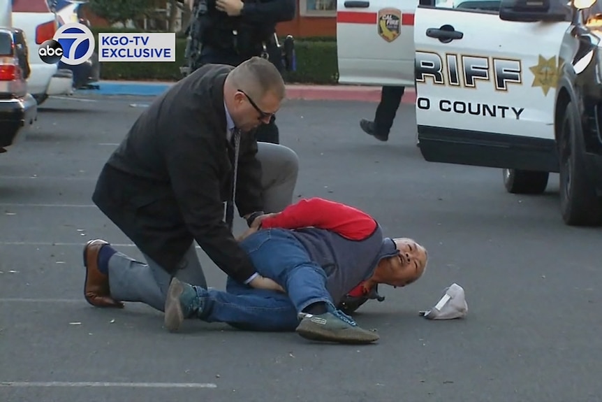 A police officer holds an older man to the ground in front of a police car. 