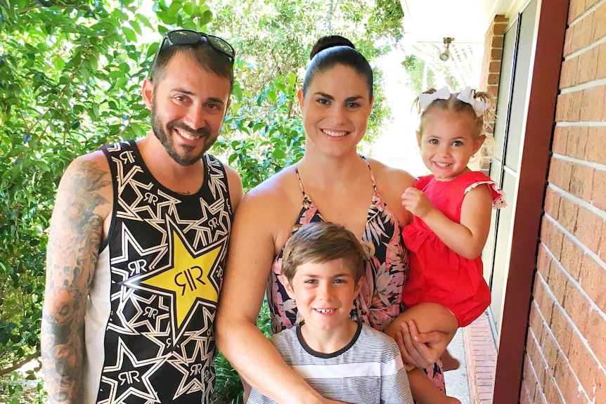 A man and woman and their daughter and son pose together for a family Christmas photo in the backyard.