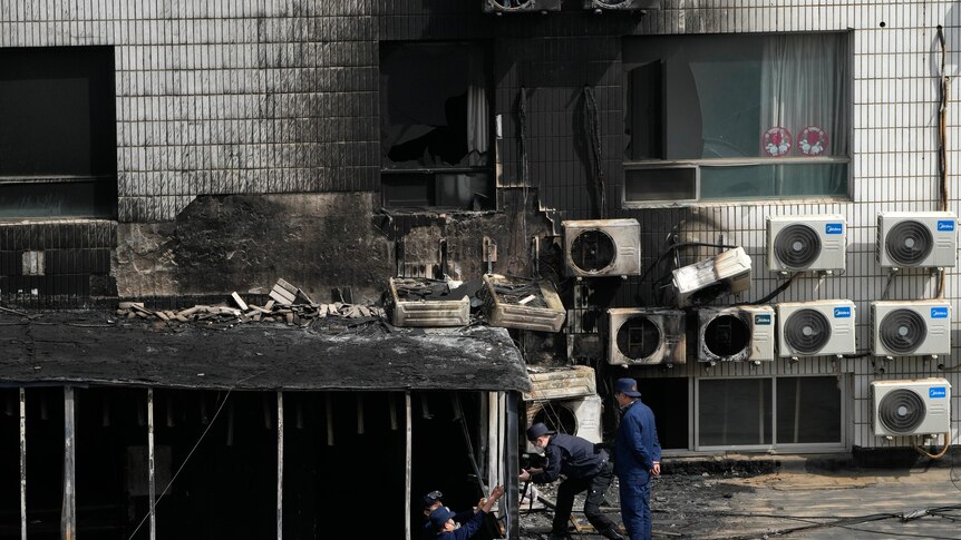Men stand next to the side of a builing that is black from fire and a number of air conditioning units.