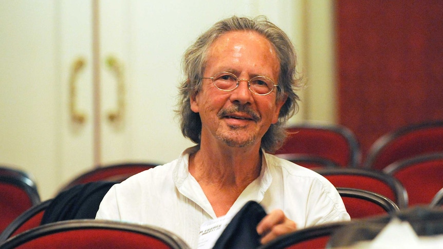 Peter Handke, wearing glasses and a white shirt and with greying long hair, smiles at the camera