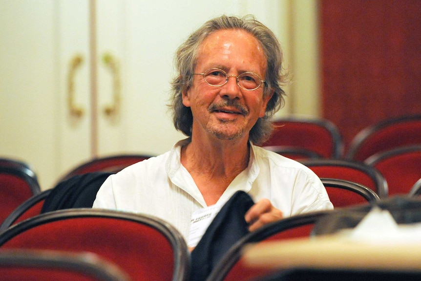 Peter Handke, wearing glasses and a white shirt and with greying long hair, smiles at the camera