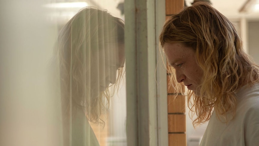 An unsmiling long-haired blond man reflected in a window.