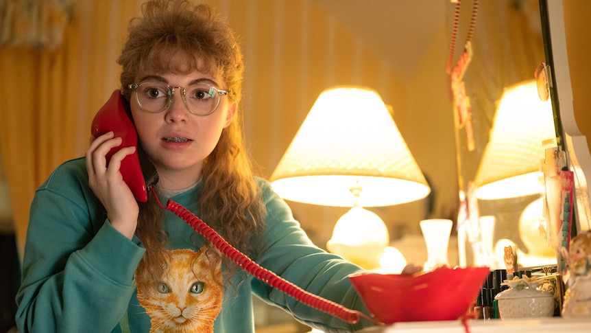 A teenage girl with a blonde mullet wears an ugly jumper and talks on a corded phone.