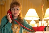 A teenage girl with a blonde mullet wears an ugly jumper and talks on a corded phone.