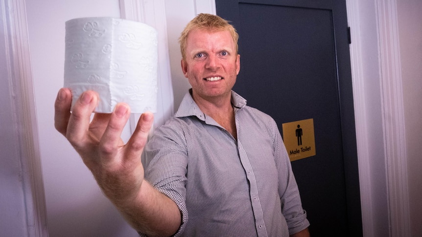 A man holds a toilet roll next to a men's toilet