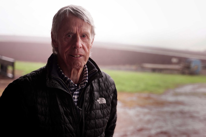 A grey-haired man in a black coat stands on a muddy farm.