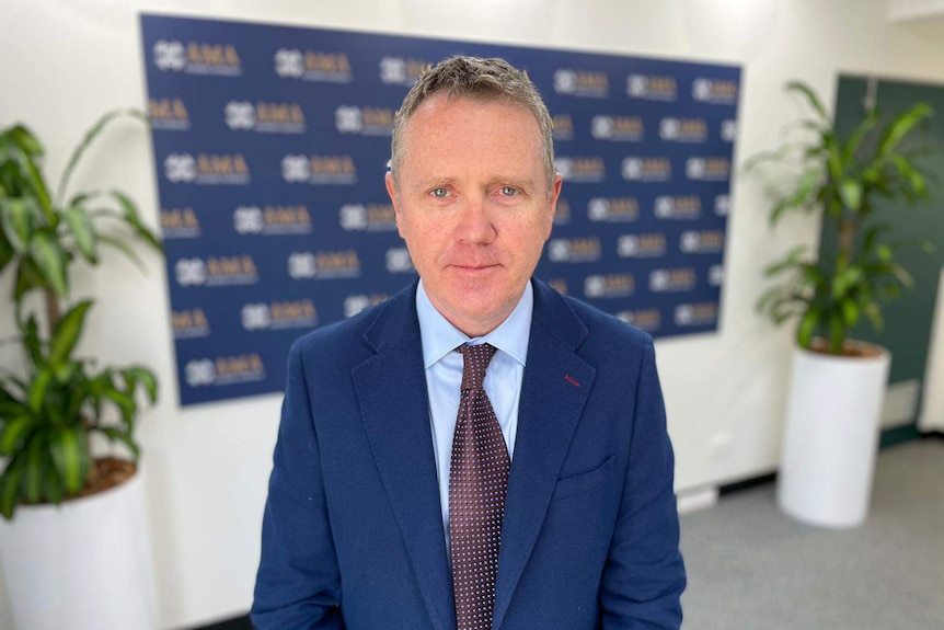 A man standing in a conference room, dressed in a dark blue suit.