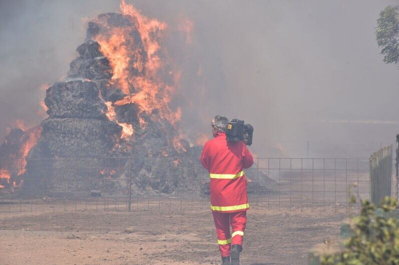 Back of cameraman Greg Ashman in red protective fire suit filming a large hay bale on fire.