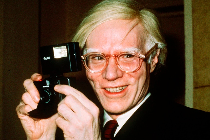 A man with blonde hair holding a photography camera smiles as he is being photographed.