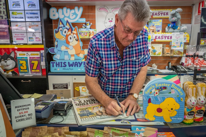 A man stands behind a counter and writes a customer's name on a newspaper.