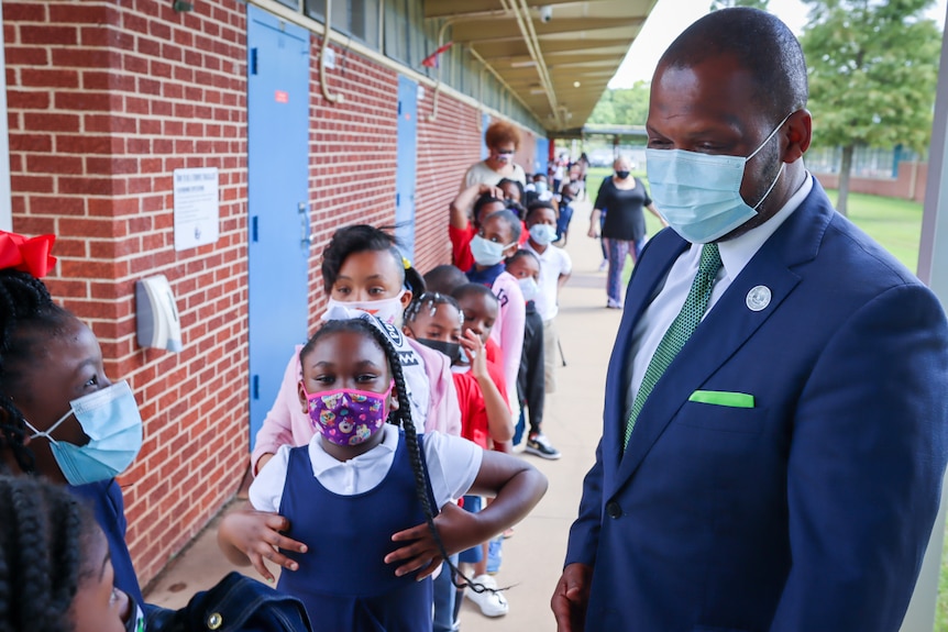 A man in a suit wearing a face mask chats with children, also masked 