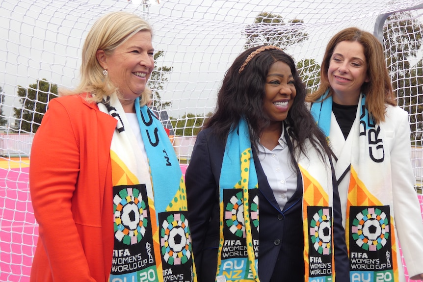 Three women wearing scarves stand in front of a football net