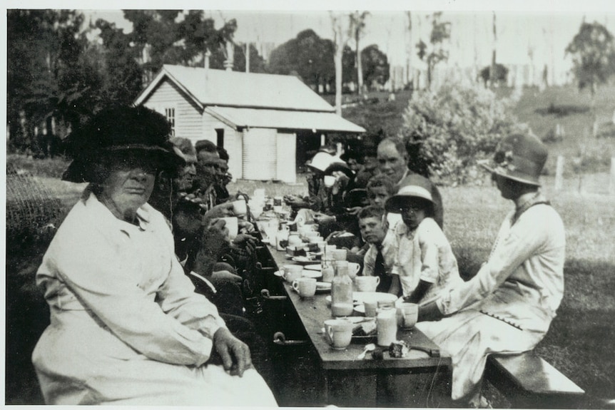 A black and white photo showing children and adults eating at an outdoor table.