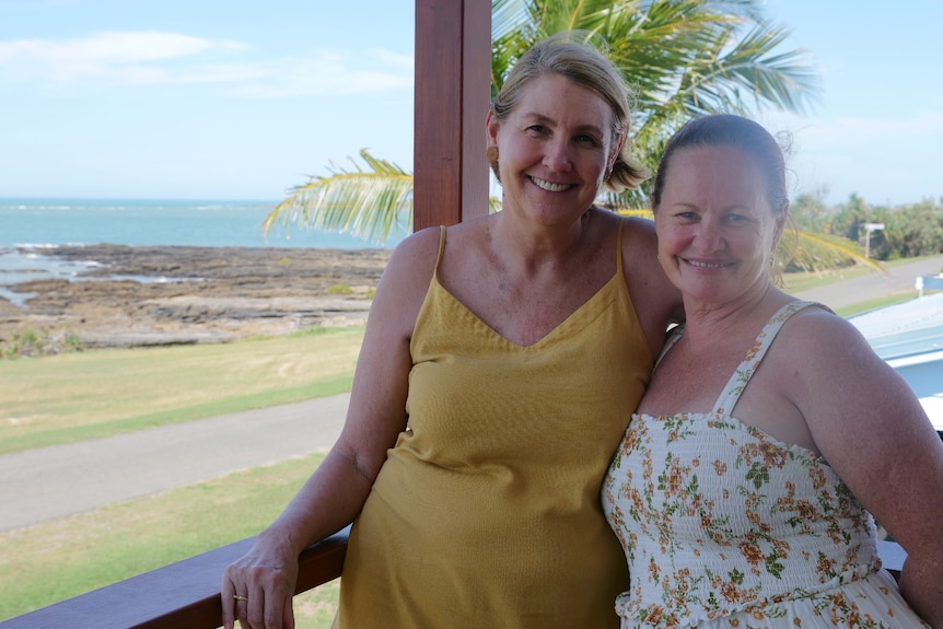Two ladies smiling, arms around each other, palm tree, ocean and rocks behind.