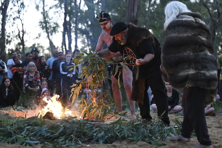 A group of people, including Indigenous, gathered around a smoking ceremony in a clearing, trees around it.