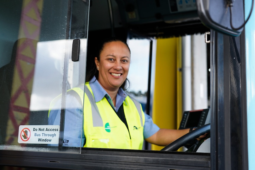 A female bus driver inside a bus holding onto the steering wheel as she smiles at the camera