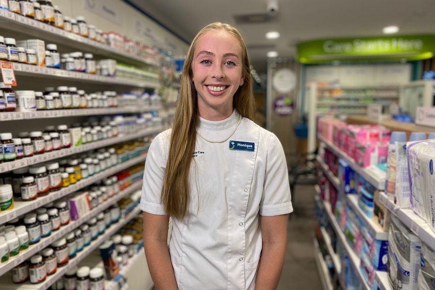 A woman with long blonde hair wearing a white pharmacist's coat stands next to shelves of medication.