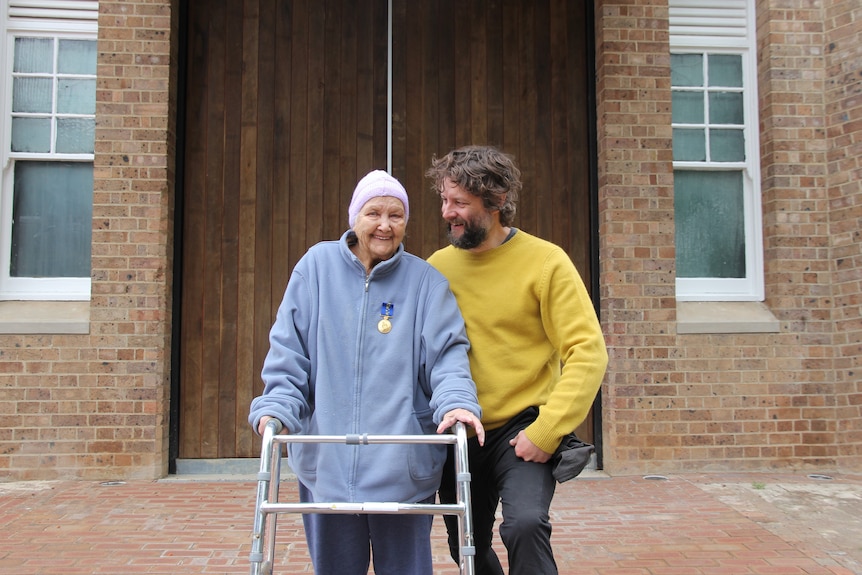 Aunty Val smiles while holding a walking frame, Ben Quilty looks at her wearing a yellow jumper.