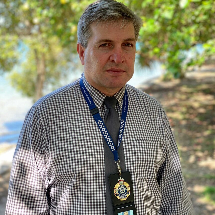 A serious police detective with his badge around his neck stands with blurred trees in the background.