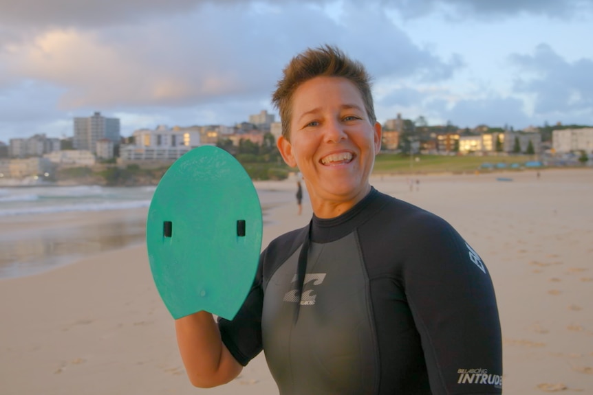 Woman in wetsuit on a beach, smiling and holding a flat piece of green plastic in her hand.