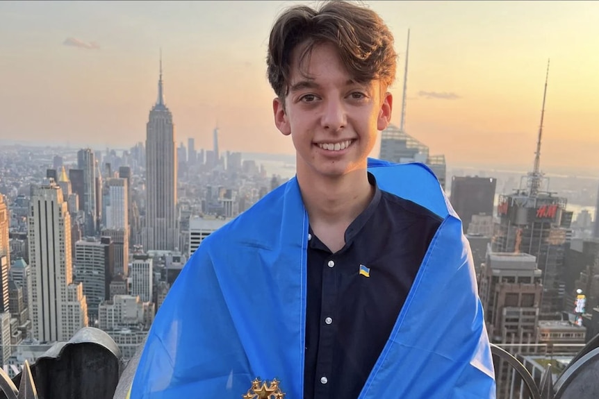 A teenage boy stands in front of the New York skyline with a Ukrainian flag around his shoulders. 