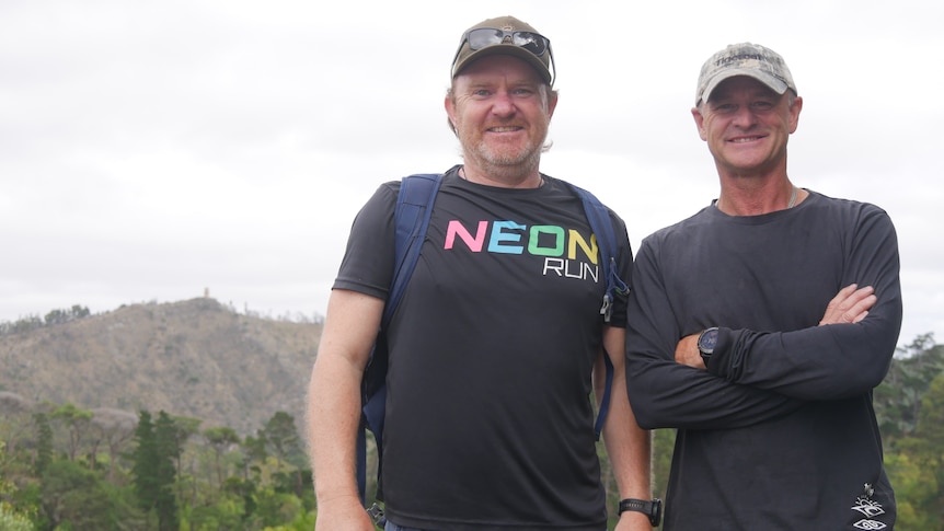 Two men smiling for the camera, one wearing a back pack, with trees and a hill in the distance