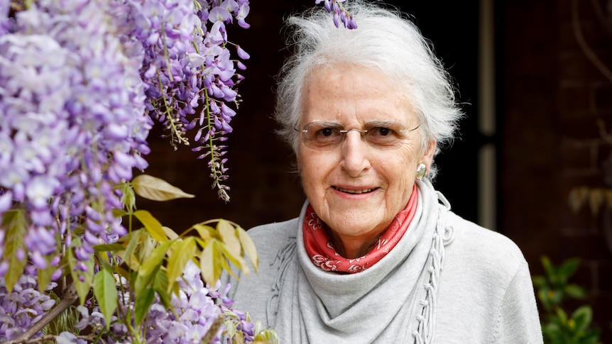 A smiling, older woman stands next a blossoming wisteria tree.